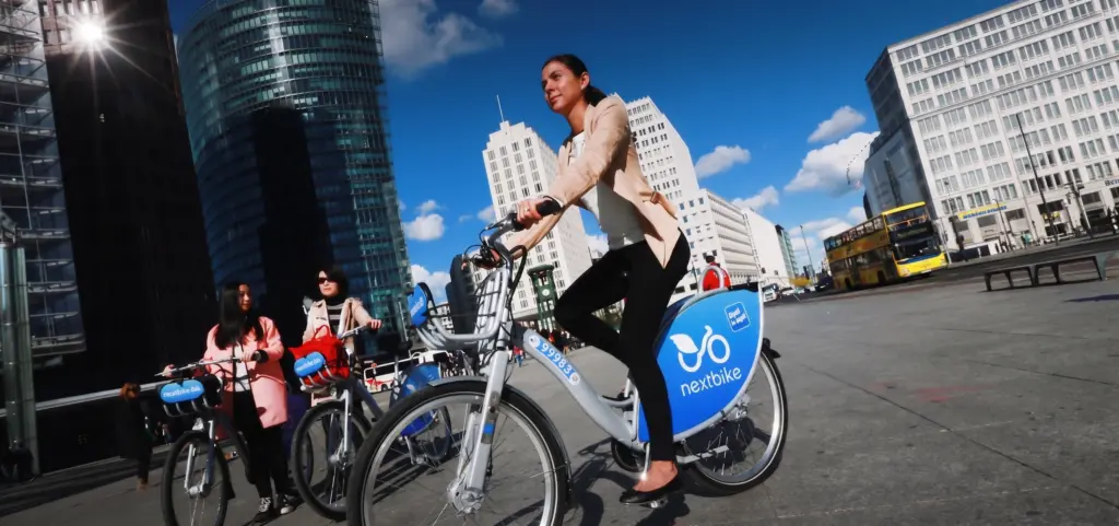 Eine Frau fährt auf einem Nextbike-Leihfahrrad in Berlin, während im Hintergrund zwei weitere Personen mit Nextbikes am Potsdamer Platz stehen.