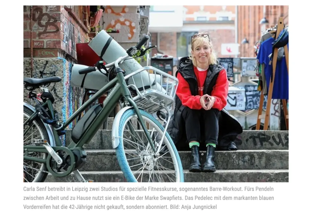 Editorial-Porträt von Anja Jungnickel, veröffentlicht in der Sächsischen Zeitung. Eine Frau sitzt lächelnd auf Treppenstufen vor einer urbanen, mit Graffiti verzierten Hauswand in Leipzig. Im Vordergrund steht ein grünes Swapfiets E-Bike mit blauem Vorderreifen und einer Yogamatte auf dem Gepäckträger. Rechts im Hintergrund befindet sich ein Kleiderständer. Unter dem Bild steht die Bildunterschrift über Carla Senf und der Fotonachweis "Bild: Anja Jungnickel".