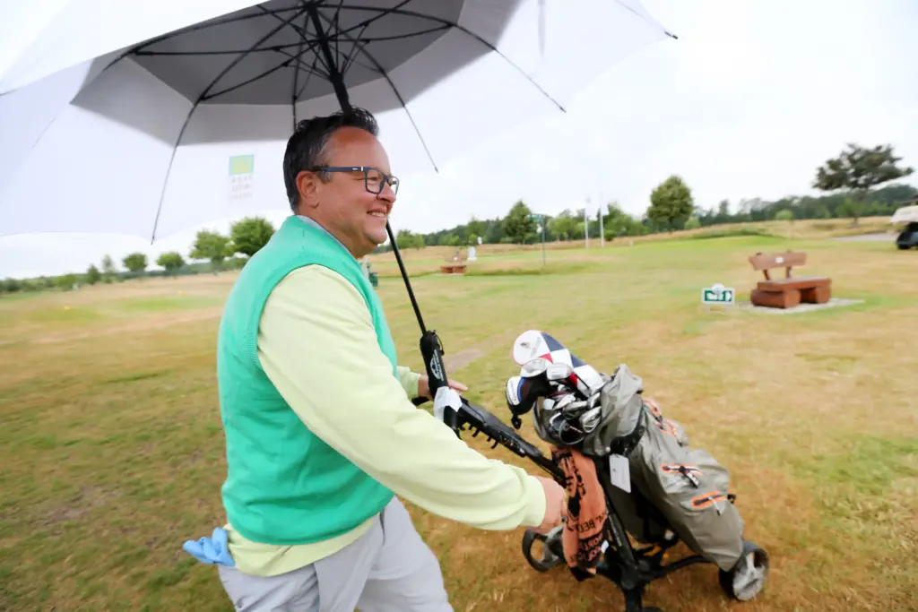 Professionelle Unternehmens- und Eventfotografie von Anja Jungnickel beim Porsche Golf Cup in Machern bei Leipzig: Ein lächelnder Golfer in grünem Pullover zieht seinen Golftrolley unter einem Regenschirm[cite: 22].